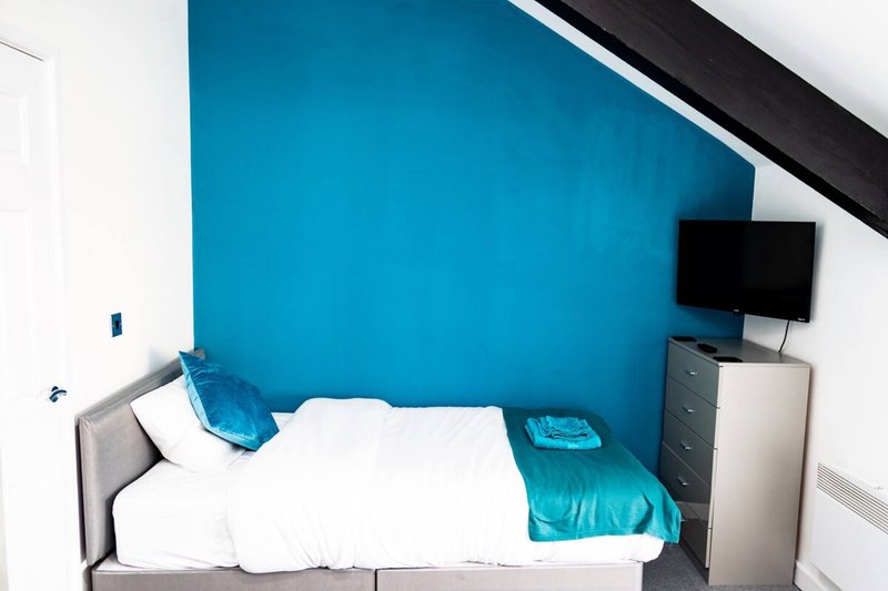 Bedroom with blue accent wall, single bed, white bedding, turquoise cushions, wall-mounted TV