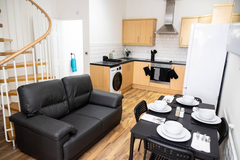 Open-plan kitchen and living area with black sofa, dining table, wooden cabinets and stairs