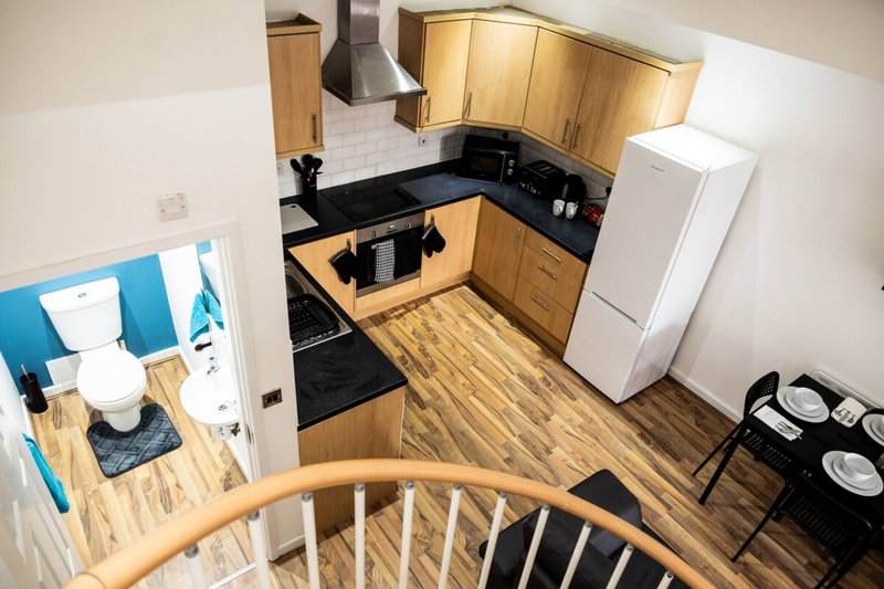 Kitchen with wooden cabinets, black countertops, white fridge, and wooden flooring viewed from stairs
