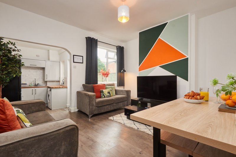 Open-plan living area with grey sofas, wooden dining table, geometric wall art, and kitchen visible in background