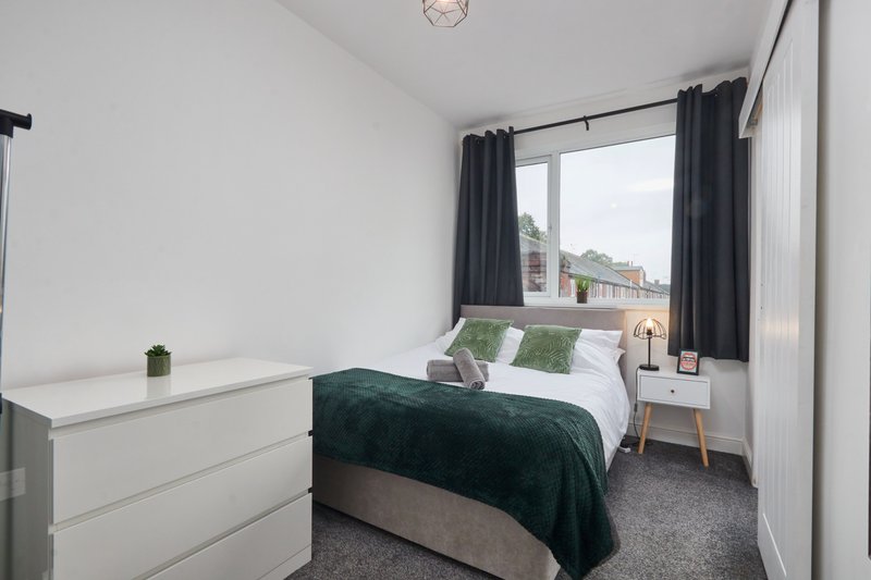 Modern bedroom with double bed, dark green throw, white dresser and window with garden view