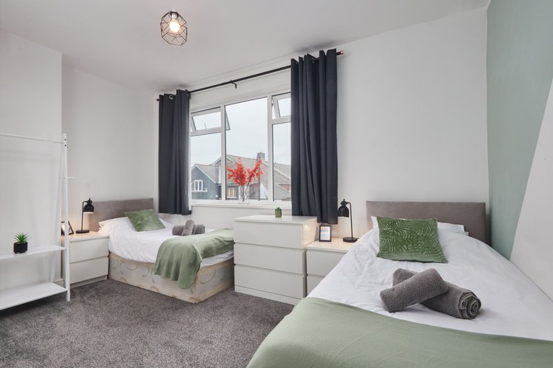 Bright bedroom with two beds, green bedding, grey carpet, and large window with dark curtains