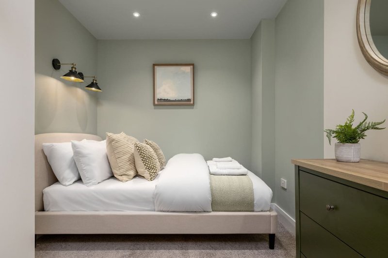 Modern bedroom with sage green walls, white bedding, decorative pillows and potted plant on dresser