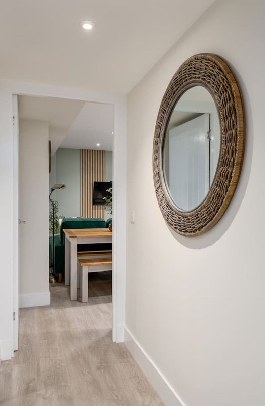 Hallway with white walls, wooden mirror, open doorway to furnished room beyond
