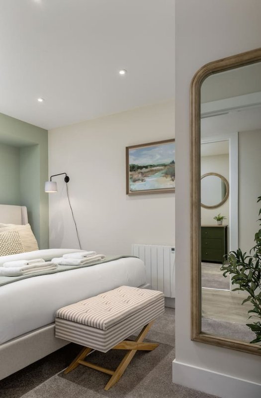 Modern bedroom with white bed, striped ottoman, wooden-framed mirror and framed landscape artwork