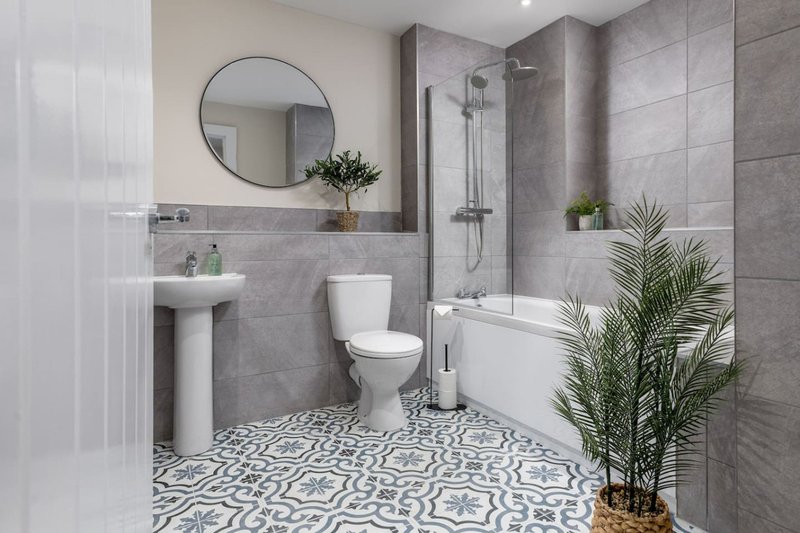 Modern bathroom with grey tile walls, patterned floor, pedestal sink, toilet and shower enclosure