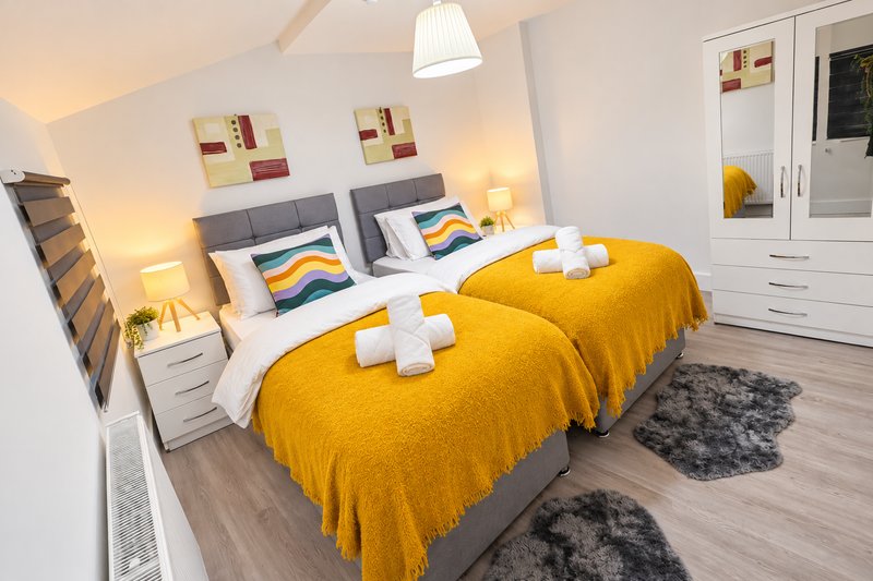 Modern bedroom with two twin beds, yellow bedding, white nightstands, pendant light, and wooden flooring