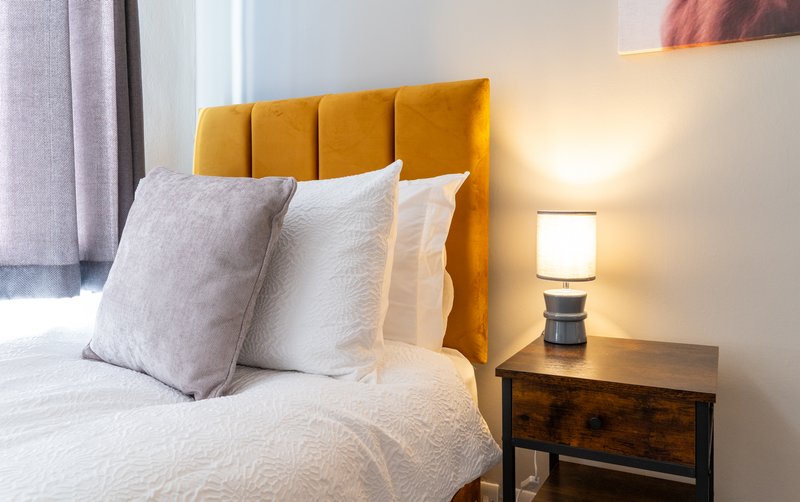 Modern bedroom with mustard yellow headboard, white bedding, decorative pillows and bedside lamp