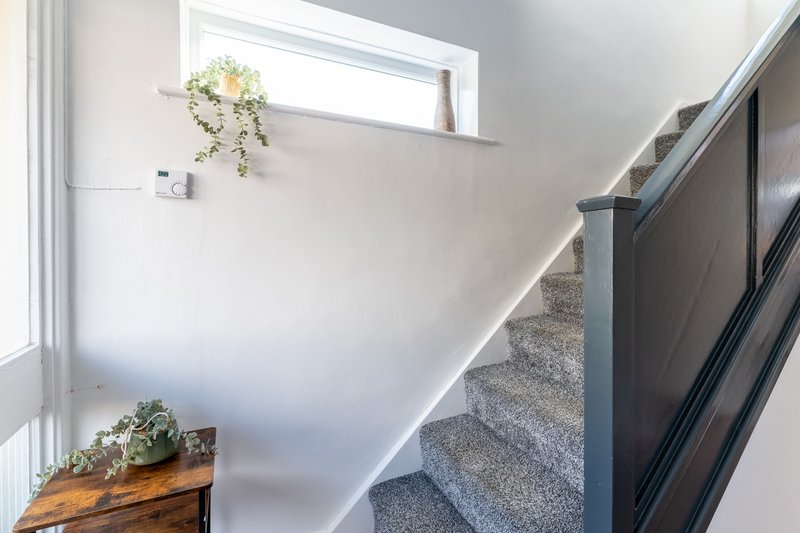 Bright stairwell with gray carpet, white walls, wooden table with plant, and hanging flowers
