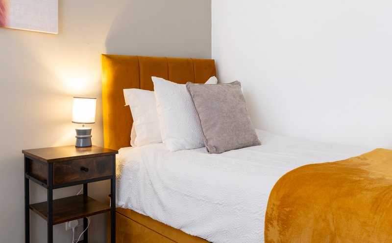 Bedroom with mustard yellow headboard, white bedding, gray pillows, bedside lamp and wooden nightstand