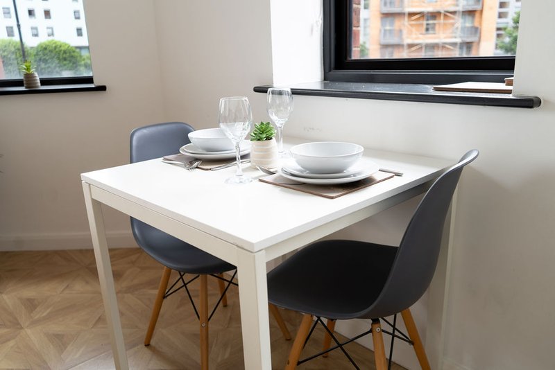 Modern dining table set for two with black chairs, place settings, and windows overlooking residential buildings
