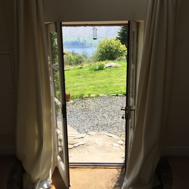Open glass door with curtains framing garden, gravel path, lawn and water view beyond