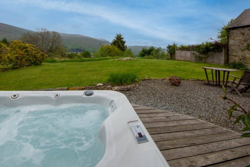 Hot tub on wooden deck overlooking green garden with mountains and stone buildings
