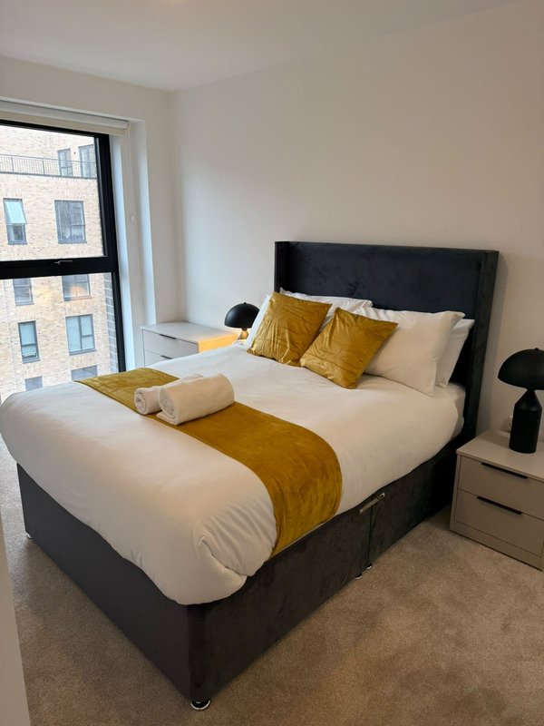 Modern bedroom with double bed, gold and cream bedding, black headboard, and city views through window
