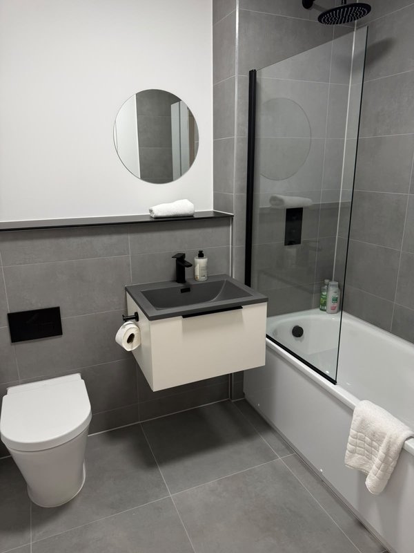 Modern bathroom with gray tile, white vanity, round mirror, toilet, and shower enclosure