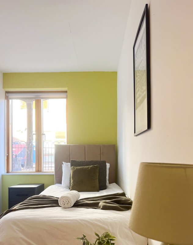 Compact bedroom with single bed, olive headboard, yellow-green accent wall, and glass door to balcony