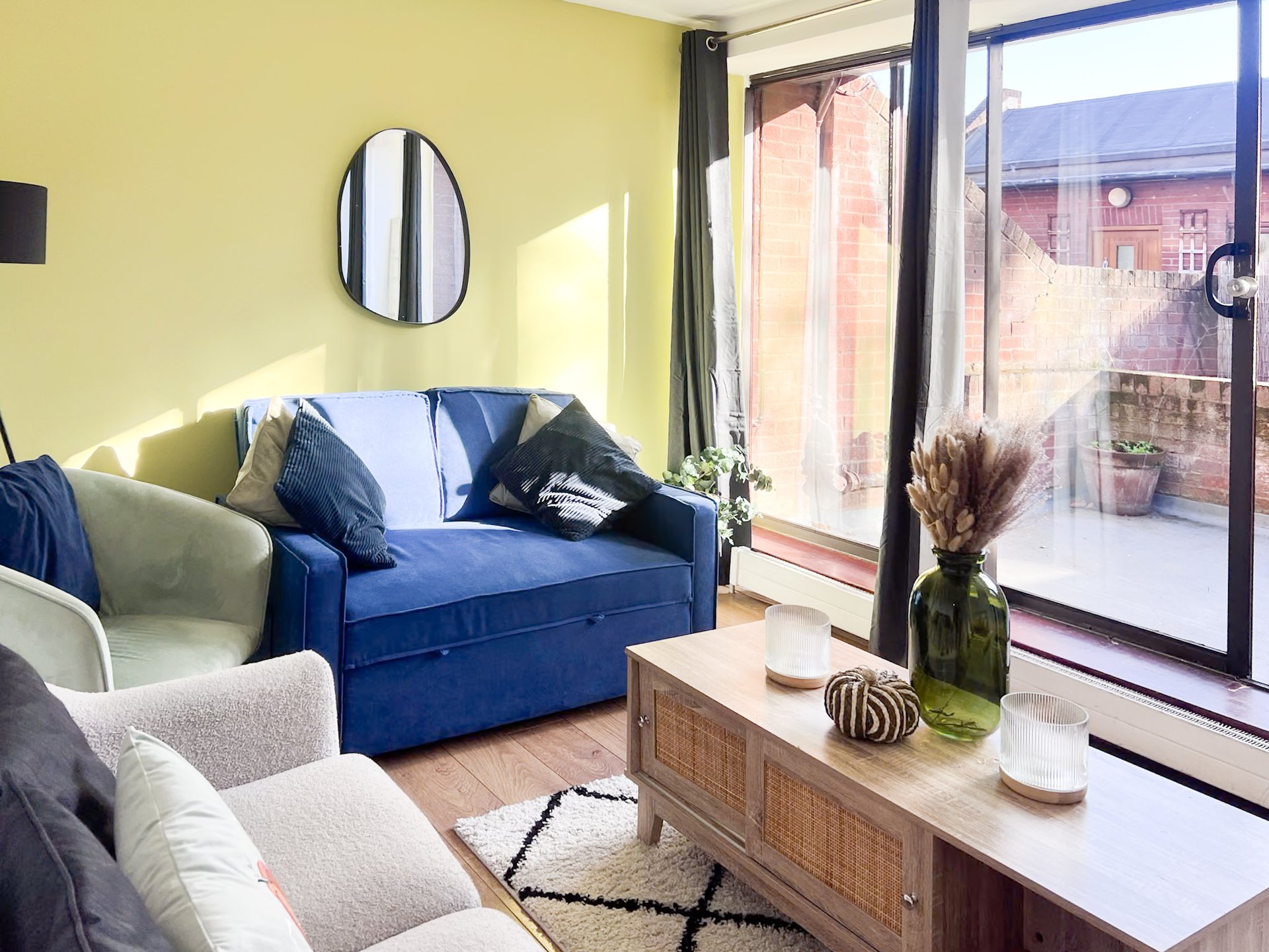 Bright living room with blue sofa, cream armchair, wooden table, and large windows overlooking courtyard