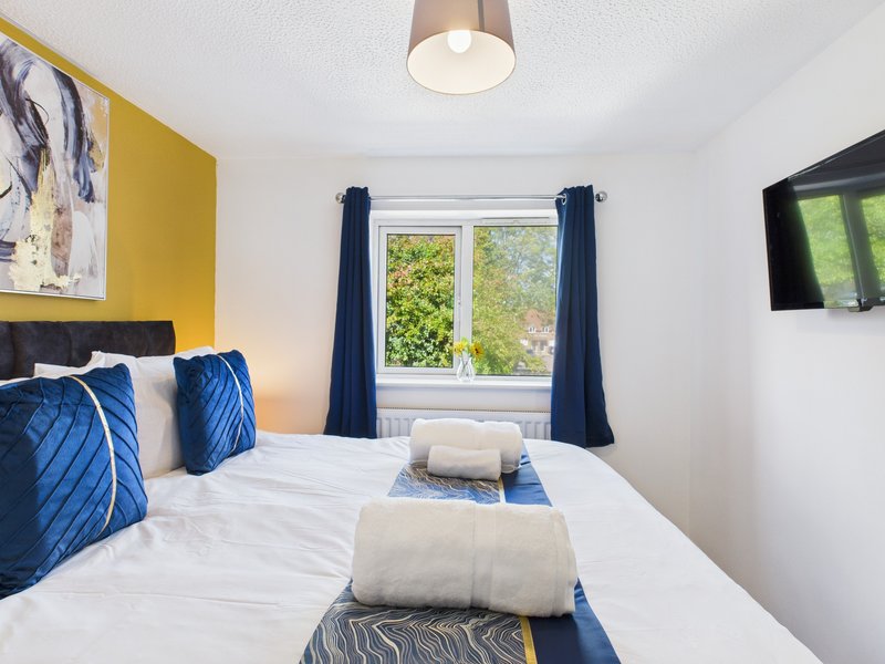 Bright bedroom with double bed, blue cushions, white bedding, yellow accent wall and window
