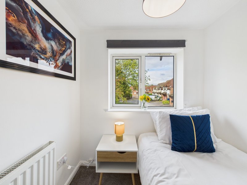 Bright bedroom with single bed, white bedding, blue cushion, small wooden nightstand and window overlooking garden
