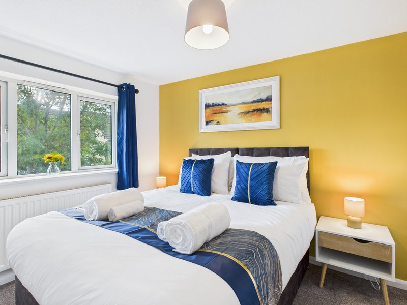 Bright bedroom with yellow accent wall, double bed with blue and white bedding, and window with garden view
