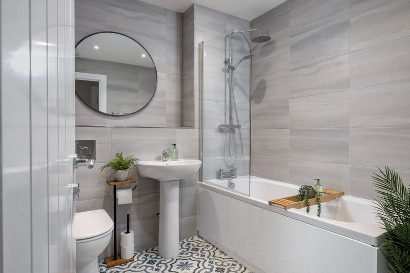Modern bathroom with white suite, grey tile walls, round mirror, and bathtub with shower screen