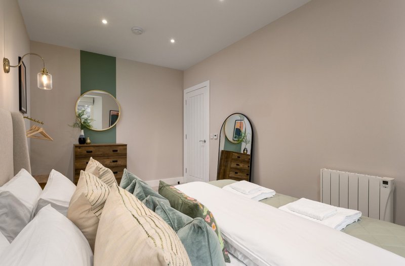 Bedroom with double bed, white bedding, green accent wall, wooden dresser and round mirror