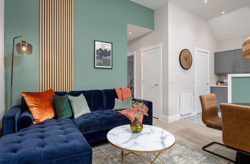 Modern living room with blue velvet sectional sofa, colorful cushions, marble-top coffee table, and teal accent wall