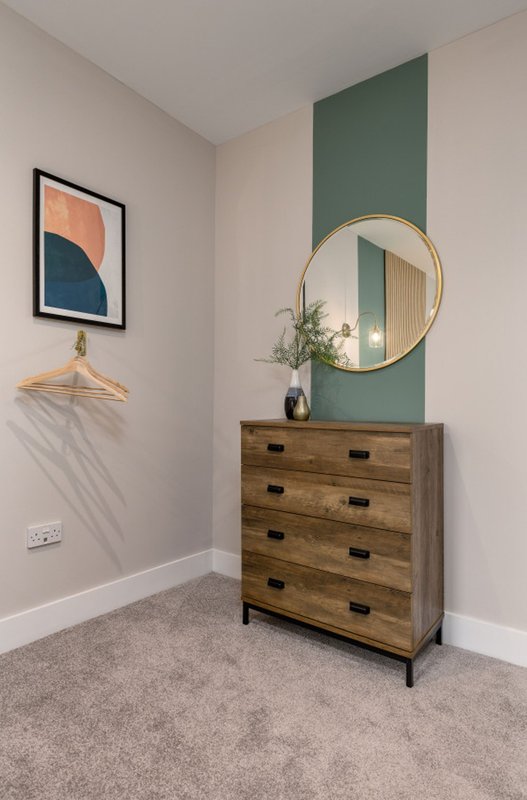 Bedroom corner with wooden chest of drawers, round mirror, framed artwork and green accent wall