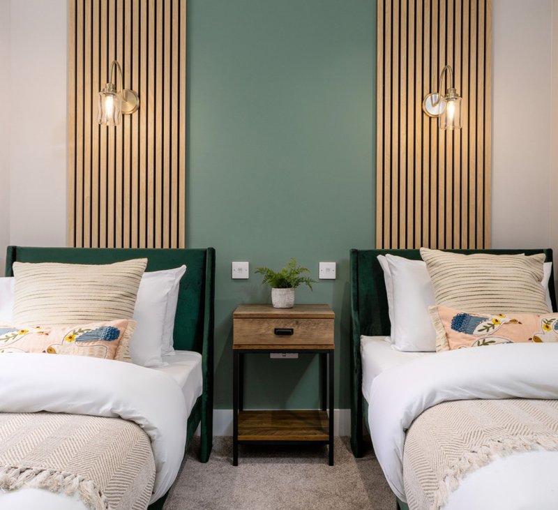 Twin bedroom with green upholstered headboards, wooden side table, pendant lights and teal accent wall