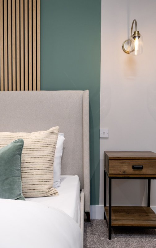 Modern bedroom with grey upholstered headboard, green and cream pillows, wooden bedside table, and wall sconce lighting