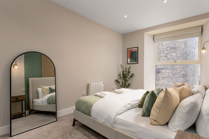 Bedroom with double bed, green and beige cushions, arched mirror, and window with stone view
