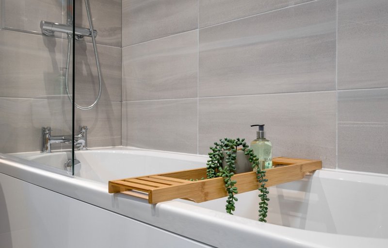 Modern bathroom with white bathtub, wooden caddy, potted plant and tiled walls