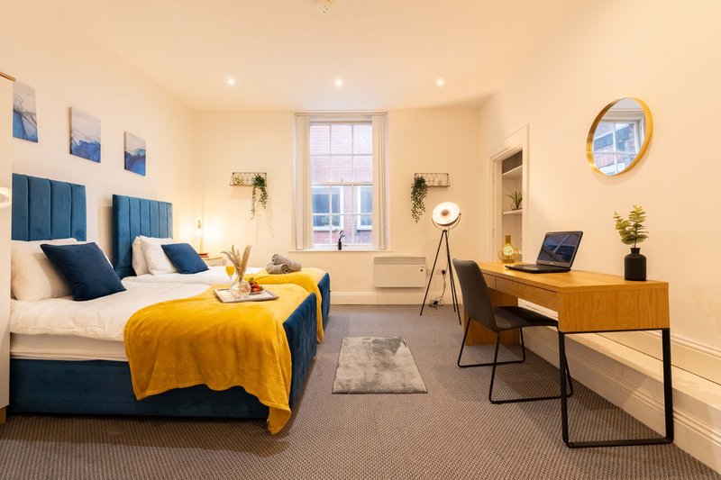 Modern bedroom with blue upholstered bed, yellow throw, desk area, and window with natural light
