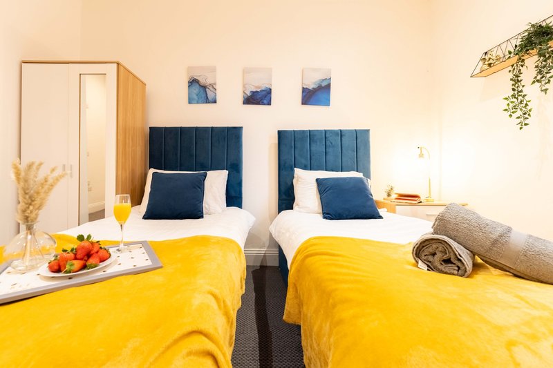 Bright twin bedroom with yellow bedding, blue headboards, and modern decor including artwork and plants