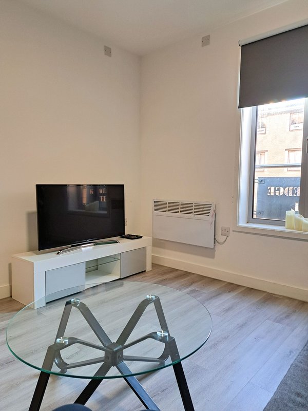 Bright living room with TV on white stand, glass table, electric heater, and window with roller blind