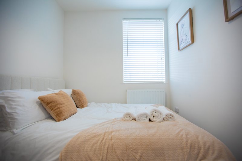 Bright bedroom with double bed, gold cushions, white bedding, and window with blinds