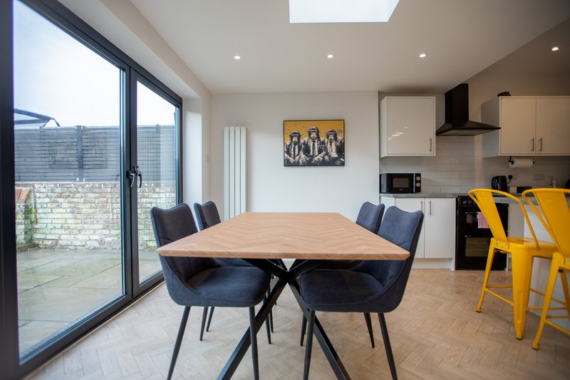 Open-plan kitchen-diner with wooden table, black chairs, yellow stools, and patio doors