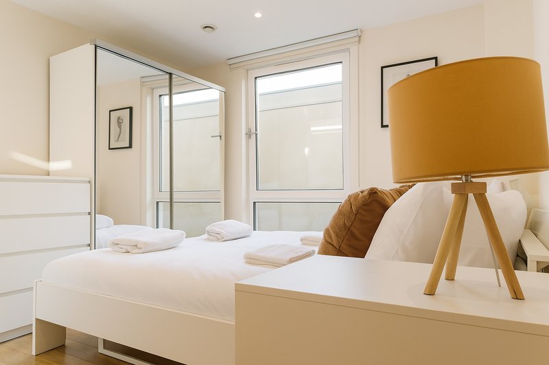 Modern bedroom with white bed, ensuite bathroom, mustard-colored tripod lamp, and large windows