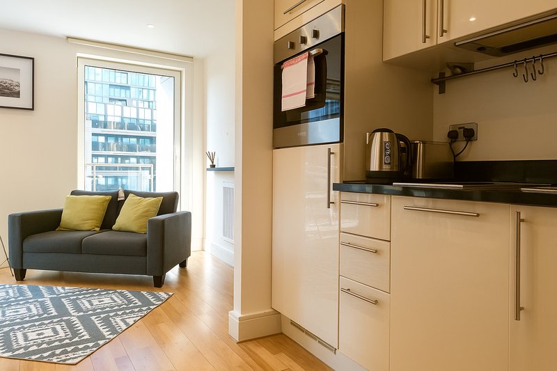 Open-plan kitchen and living area with cream cabinets, black countertop, and grey sofa with yellow cushions