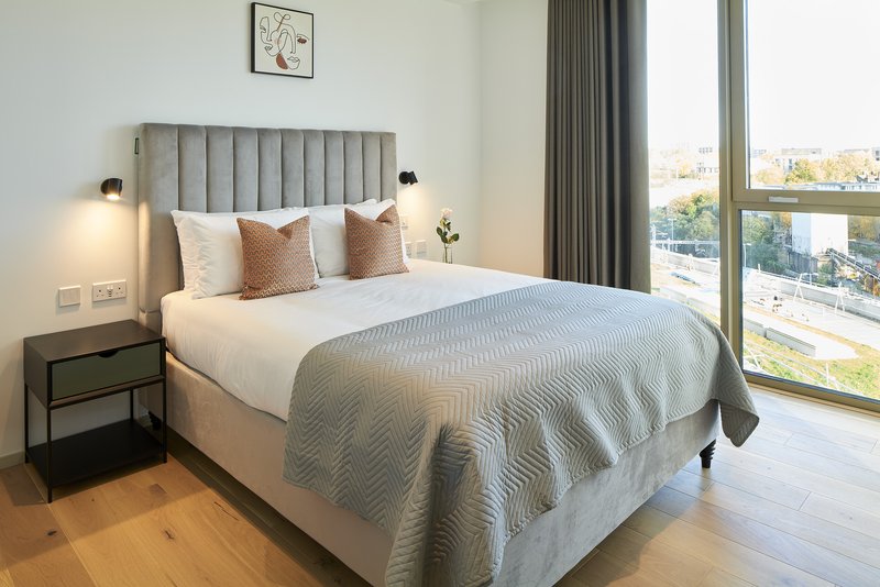 Modern bedroom with grey upholstered bed, bedside table, large windows overlooking city