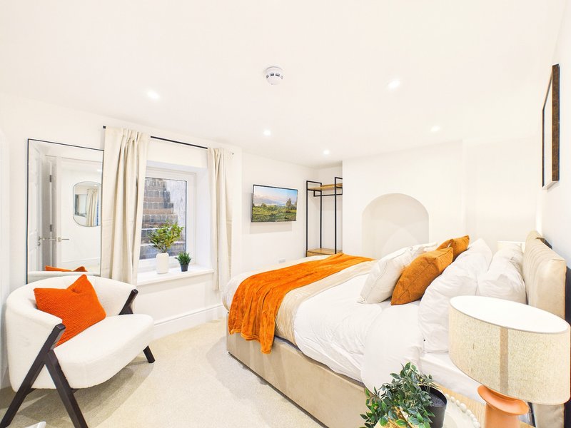 Bright modern bedroom with double bed, orange throw, white armchair and large windows overlooking garden
