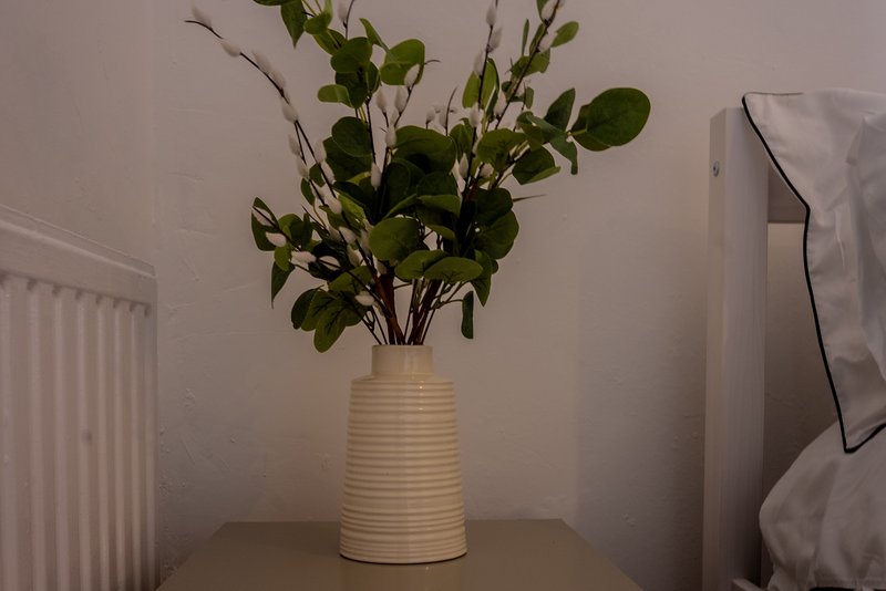 Cream ceramic vase with fresh green foliage and branches on bedside table