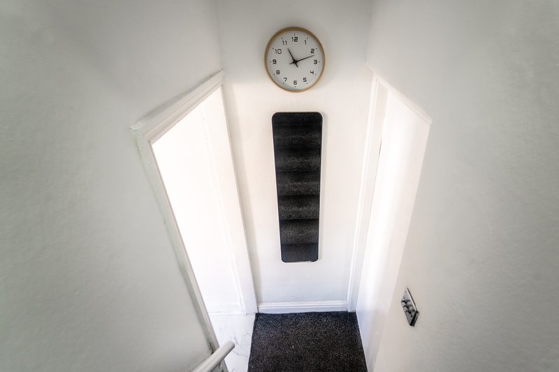 Narrow hallway with white walls, wall clock, black shoe organizer and dark runner mat