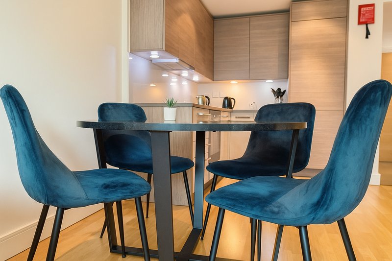 Modern dining area with round black table and four blue velvet chairs, open-plan kitchen behind