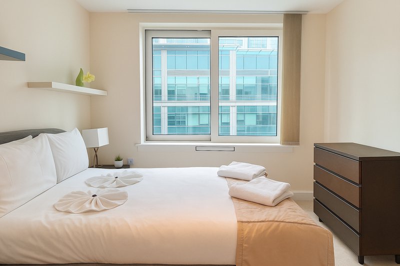 Modern bedroom with double bed, white bedding, dark wood dresser and city view windows