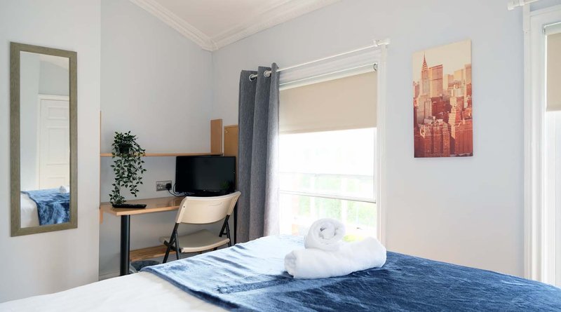 Bright bedroom with double bed, desk, chair, mirror and cityscape artwork on walls