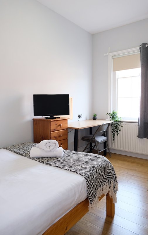 Bright bedroom with single bed, wooden TV stand, desk, chair and window with curtains