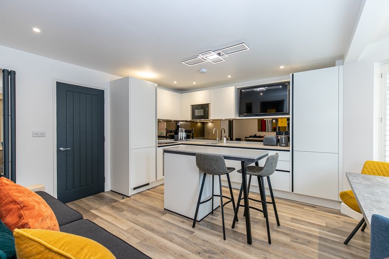 Modern open-plan kitchen and living area with white cabinetry, breakfast bar, wooden flooring, and colorful seating