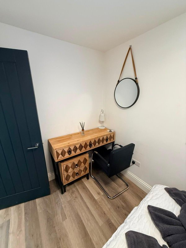 Bedroom corner with wooden dresser, round mirror, dark door, and bed with white and black bedding