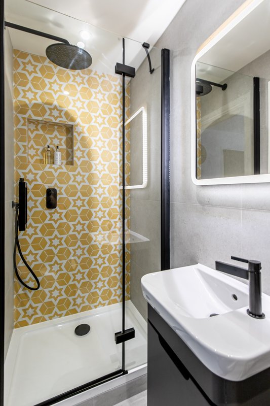 Modern bathroom with yellow geometric tile shower, glass enclosure, white sink and illuminated mirror